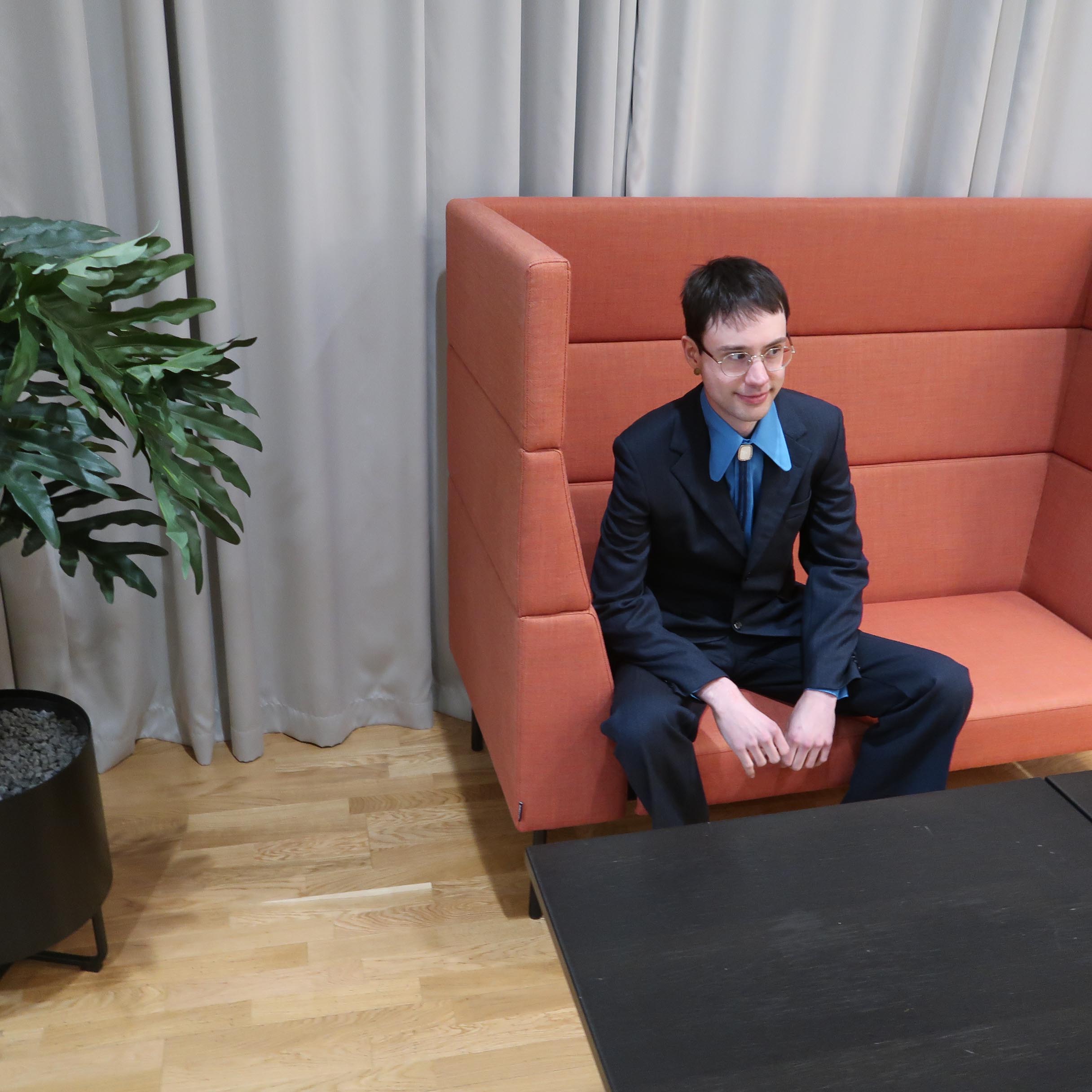 Ian on an orange couch in a spiffy suit by a fake plant.
